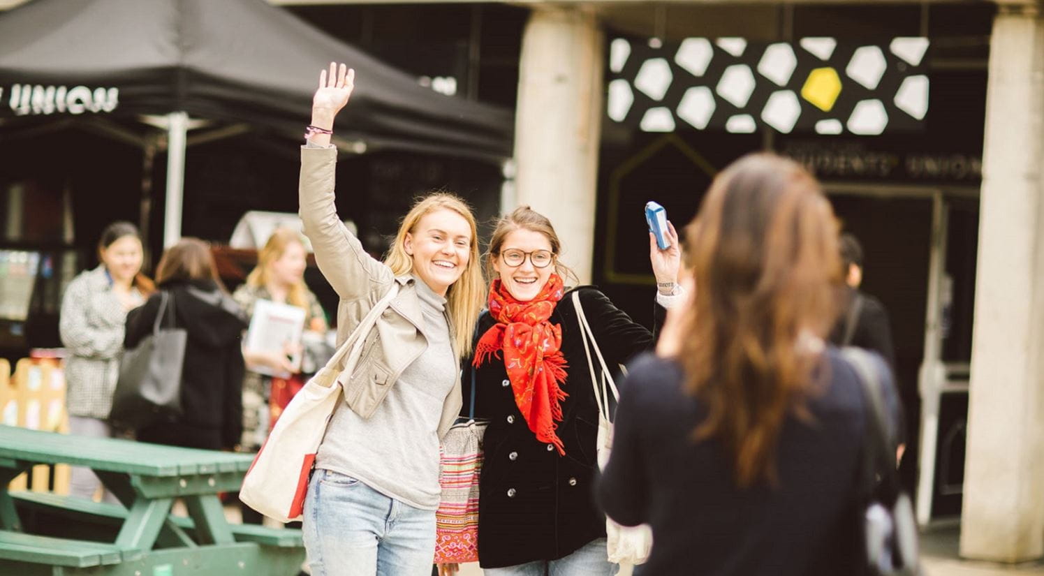 Two students standing next to each other outside the Students' Union with arms raised having their photo taken)